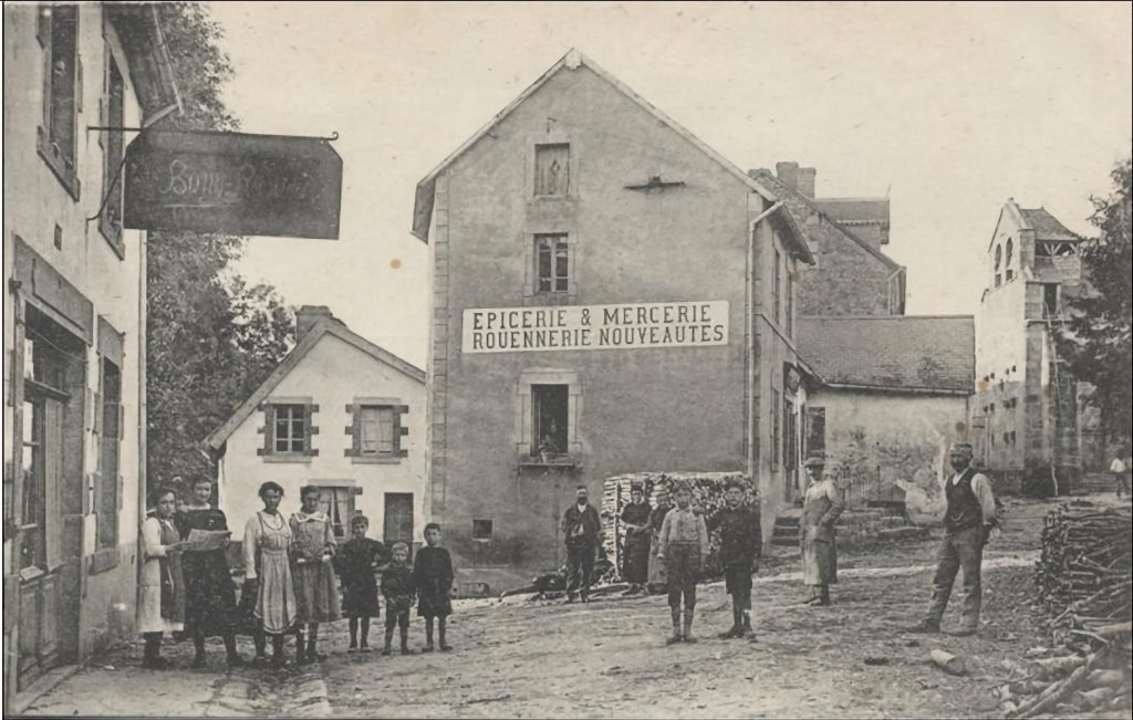 Photographie ancienne en noir et blanc du village de Saint-Avit, montrant une épicerie-mercerie avec une enseigne "Rouennerie Nouveautés". Plusieurs habitants, dont des enfants et des adultes en tenue d'époque, posent devant les bâtiments. Une église est visible en arrière-plan, ajoutant au charme historique de la scène.
