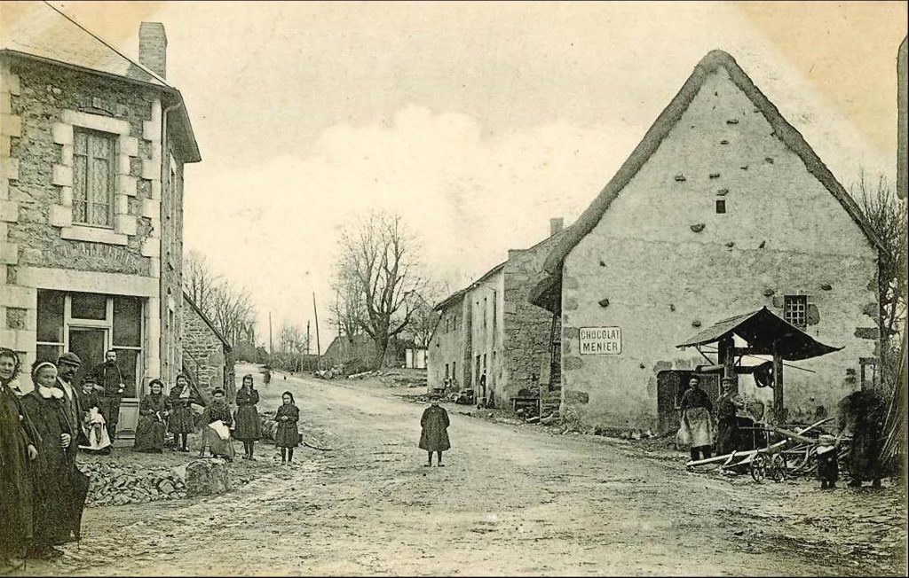 Photographie ancienne en noir et blanc du village de Saint-Avit. Une rue en terre traverse l’image, bordée de maisons en pierre et en torchis. À gauche, un groupe d’habitants, principalement des femmes et des enfants, posent devant une bâtisse en pierre. À droite, une maison au toit de chaume affiche une enseigne "Chocolat Menier". Sous un petit auvent, des villageois semblent occupés à diverses tâches, tandis qu’un enfant se tient au milieu de la rue, ajoutant de la vie à cette scène d’époque.