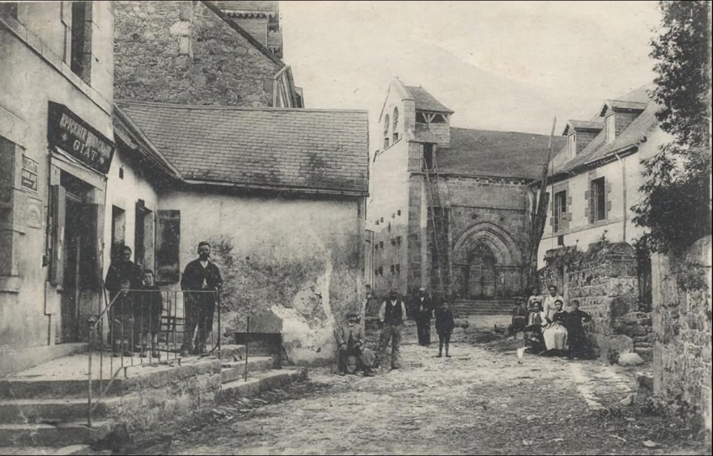 Photographie ancienne en noir et blanc du village de Saint-Avit. On y voit une épicerie-buvette avec une enseigne indiquant "Épicerie Buvette GIAT", où trois hommes, dont un enfant, posent sur un perron. La rue pavée mène à une église en pierre au fond de l'image, avec son clocher et son portail roman. Plusieurs habitants, hommes, femmes et enfants, sont assis ou debout, donnant une atmosphère vivante à cette scène de village d'époque.